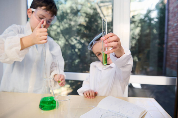 Focus selettivo su una provetta con soluzione chimica verde nelle mani della bambina offuscata, guardando la reazione chimica in corso. Bambini che imparano chimica. Laboratorio scolastico. Educazione alla conoscenza - Foto, immagini