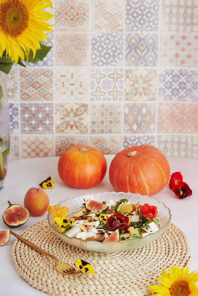 Vegetarische salade met feta, eetbare bloemen, vijgen, perzik, basilicum op keramische tegel achtergrond met zonnebloemen en pompoen decoraties - Foto, afbeelding