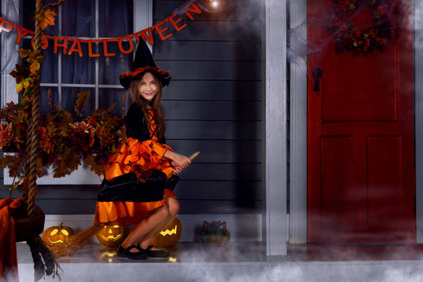 Young funny girl kid child in halloween orange costume of witch and black hat playing outdoor with witch broom and spooky jack pumpkins with scary faces, fog and autumn fail leaves. Halloween concept. - 写真・画像