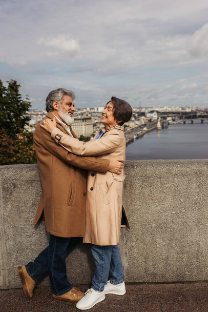 happy and senior couple in beige coats hugging on bridge near river - Photo, Image