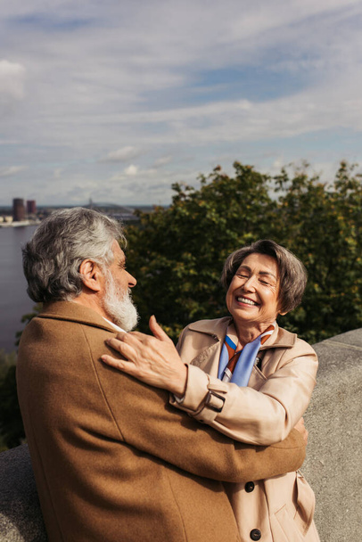 happy and senior couple in beige coats hugging each other on bridge near river - Photo, Image