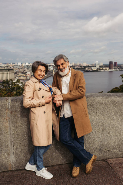 piena lunghezza di uomo anziano allegro in mano cappotto tenuta di moglie sorridente vicino al fiume in autunno  - Foto, immagini