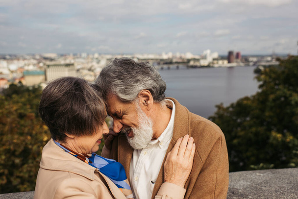 mujer mayor abrazándose con el marido barbudo con el pelo gris y sonriendo cerca del río - Foto, Imagen