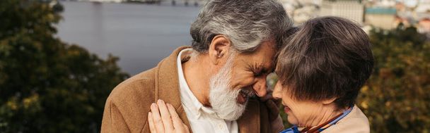 donna anziana che abbraccia con marito barbuto con i capelli grigi e sorridente vicino al fiume, striscione - Foto, immagini