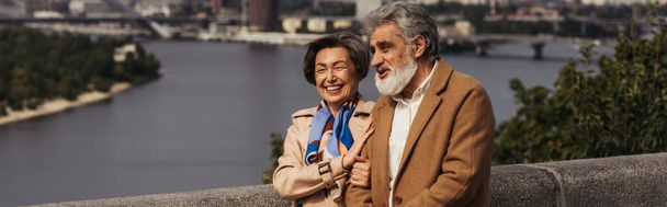 ευτυχισμένο ζευγάρι ηλικιωμένων σε μπεζ παλτά χαμογελώντας και περπατώντας στη γέφυρα κοντά στο ποτάμι, πανό  - Φωτογραφία, εικόνα