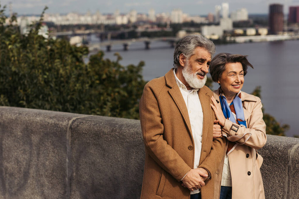 cheerful senior couple in beige coats smiling and walking on bridge near river  - Фото, изображение