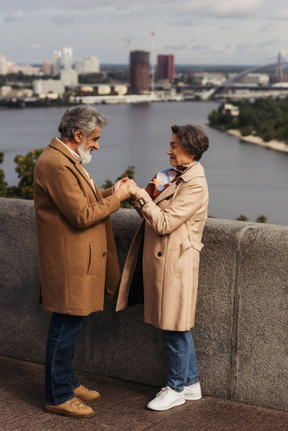 Seitenansicht eines fröhlichen Seniorehepaares in beigefarbenen Mänteln, das Händchen haltend auf einer Brücke in Flussnähe steht  - Foto, Bild