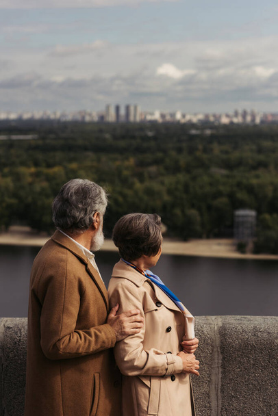 back view of senior couple in beige coats standing on bridge near river - Photo, Image