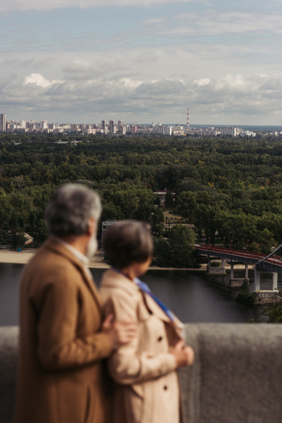 vue arrière du couple âgé flou en manteaux beiges debout sur un pont près de la rivière - Photo, image
