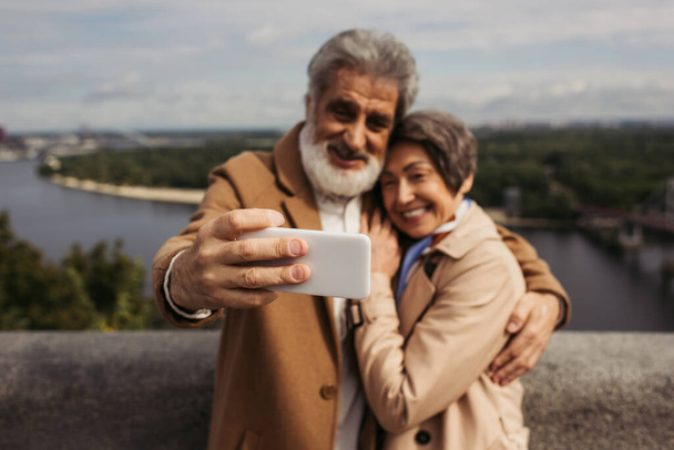 happy senior man taking selfie with cheerful wife in trench coat on focused smartphone - Photo, Image