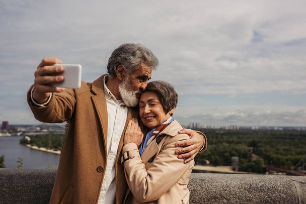 happy senior man kissing head of cheerful wife in trench coat and taking selfie - Zdjęcie, obraz