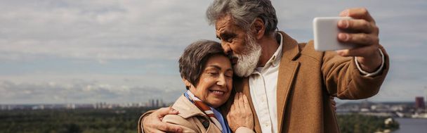 bärtiger älterer Mann küsst Kopf von fröhlicher Frau und macht Selfie, Banner - Foto, Bild