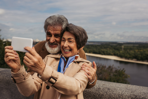 Χαρούμενη ηλικιωμένη γυναίκα με καμπαρντίνα και λήψη selfie με γενειοφόρο σύζυγο  - Φωτογραφία, εικόνα