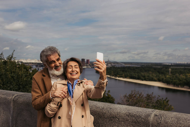 happy senior woman in beige trench coat and taking selfie with bearded husband  - Foto, imagen