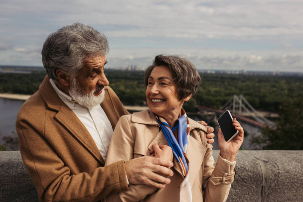 happy senior woman in beige trench coat looking at bearded husband and holding smartphone with blank screen - Photo, Image