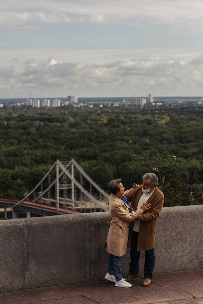 wesoła para seniorów w beżowych jesiennych płaszczach przytulających się na moście nad rzeką  - Zdjęcie, obraz