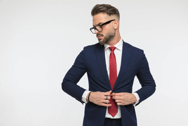 vousatý elegantní podnikatel s červenou kravatou zapínání oblek a hledá na straně na bílém pozadí ve studiu - Fotografie, Obrázek