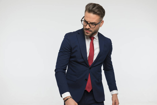 sexy homme d'affaires non rasé en costume élégant avec cravate rouge regardant loin et posant de manière confiante sur fond blanc en studio - Photo, image