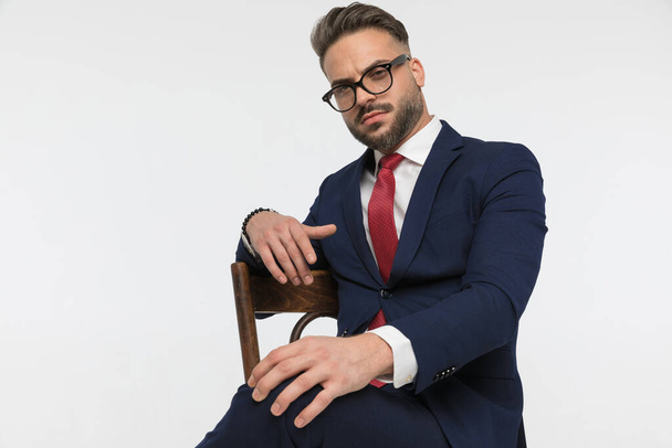 1本の腕を椅子の上に置き白い背景の前に座っている間に膝に触れるメガネを持つクールな髭のビジネスマン - 写真・画像