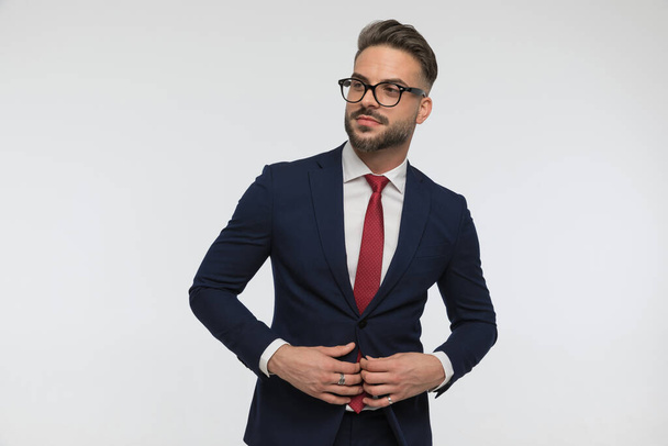 confident businessman with glasses looking away and buttoning suit in front of white background in studio - Фото, изображение