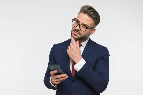 portret troskliwego biznesmena trzymającego telefon i myślącego na białym tle w studio - Zdjęcie, obraz