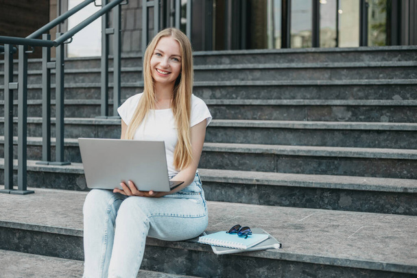 Joven estudiante rubia caucásica sentada con portátil en las escaleras cerca de la universidad sonriendo y aprender inglés. Copiar espacio - Foto, imagen