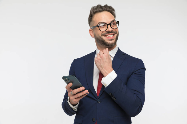 hombre de negocios feliz en traje elegante sosteniendo el teléfono, mirando hacia otro lado y sonriendo sobre fondo blanco en el estudio - Foto, imagen