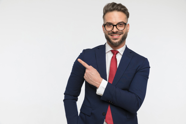gelukkig jong zakenman met bril wijzend vinger naar kant en glimlachen op witte achtergrond in studio - Foto, afbeelding