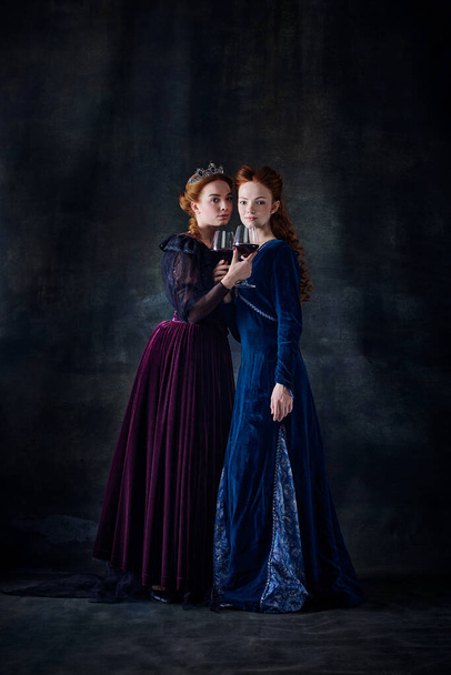 Retrato de duas mulheres bonitas na imagem de rainhas com copos de vinho isolados em fundo escuro. Celebração. Conceito de comparação de eras, modernidade e renascimento, estilo barroco, beleza, história - Foto, Imagem