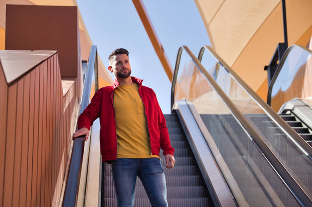 Jeune homme beau en veste rouge, t-shirt jaune et jeans descendant escaliers roulants dans un centre commercial. Concept beauté, mode, moderne, tendance. - Photo, image