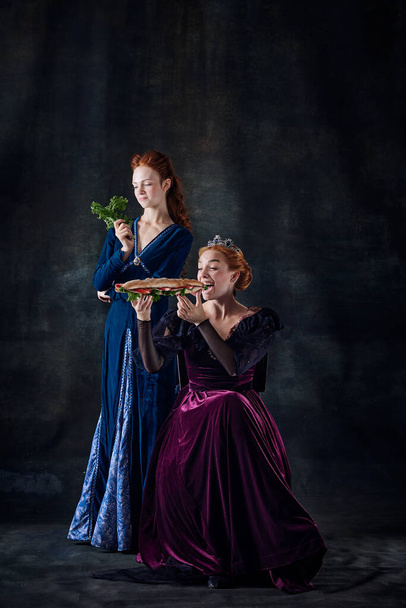 Portret van twee mooie vrouwen, koningin die broodjes eet, prinses die sla eet geïsoleerd over donkere achtergrond. Concept van vergelijking van tijdperken, moderniteit en renaissance, barokke stijl, schoonheid, geschiedenis - Foto, afbeelding