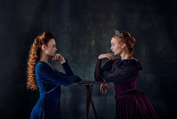 Portrait of two beautiful women in image of queens isolated over dark background. Confrontation, eye to eye. Concept of comparison of eras, modernity and renaissance, baroque style, beauty, history - Photo, Image