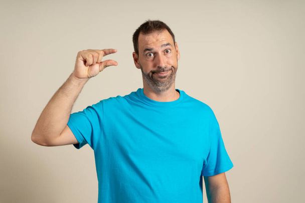 I need some more, please give me. Bearded man showing small gesture with skeptical smile, showing min sign, measuring small size. Isolated on beige studio background - Photo, Image