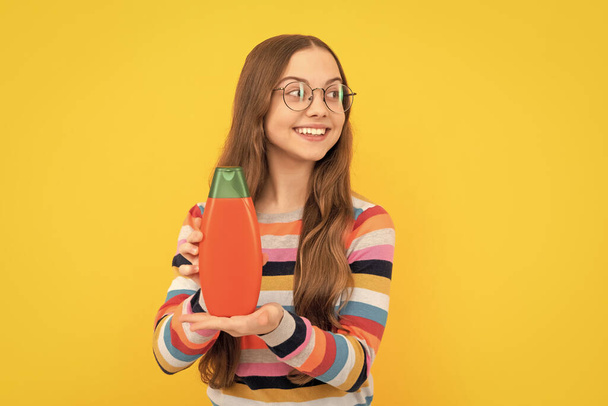 cheerful child long hair in glasses showing shower gel bottle, advertising. - Foto, afbeelding