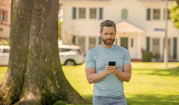hymyilevä kypsä mies seisoo ulkona talossa chattailuun puhelimessa, ostaa verkossa. - Valokuva, kuva