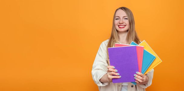 Caucasian blonde student girl holds four books in multi-colored covers smiling isolated over orange background in studio. English language school, education concept - Foto, imagen