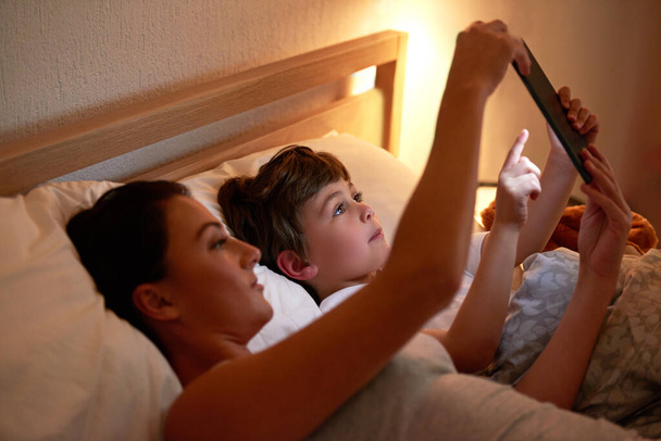 Interaktywne opowieści na dobranoc. matka i syn korzystający z tabletu cyfrowego razem leżąc w łóżku - Zdjęcie, obraz