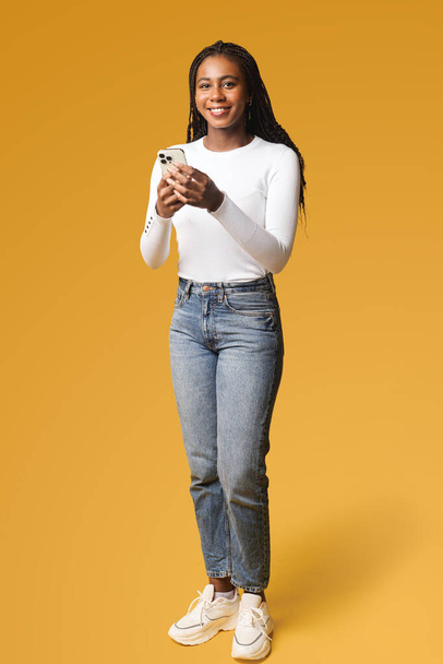 Πολυφυλετική γυναίκα που κρατά το smartphone, κουβεντιάζοντας στα κοινωνικά δίκτυα, χρησιμοποιώντας την εφαρμογή για κινητά, στέλνοντας μηνύματα στο διαδίκτυο με φίλους, κοινοποιώντας ειδήσεις, απομονωμένη στο κίτρινο - Φωτογραφία, εικόνα