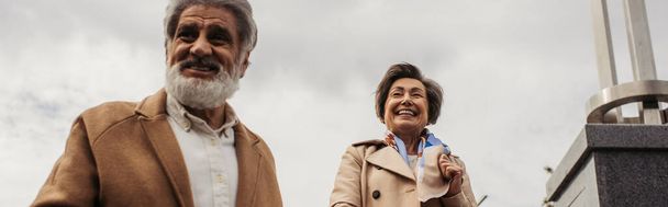 pleased senior man in coat smiling with happy wife against cloudy sky, banner - Fotó, kép