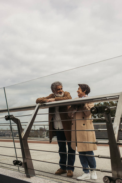 cheerful senior man in coat looking at pleased wife while leaning on guard rail of bridge  - Φωτογραφία, εικόνα