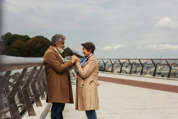 oldalnézetben boldog idős nő kéz a kézben szakállas és vidám férj közel híd őr sín  - Fotó, kép
