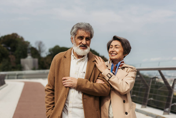Fröhliches Seniorenpaar in beigen Herbstmänteln lächelt beim Gang über Brücke - Foto, Bild