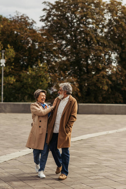 vrolijke senior vrouw knuffelen bebaarde man in jas en staan in het stadspark - Foto, afbeelding