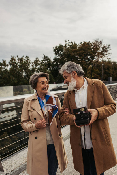 счастливый пожилой человек держит винтажную камеру возле улыбающейся жены на мосту  - Фото, изображение