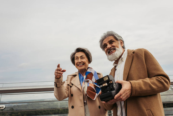 bärtiger und älterer Mann mit Vintage-Kamera neben Frau lächelnd, während er mit Finger auf Brücke zeigt  - Foto, Bild