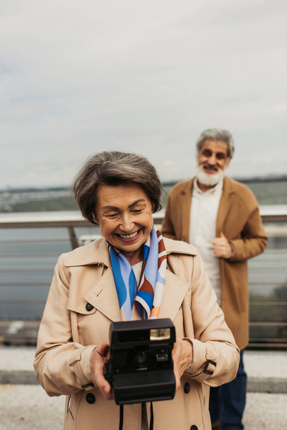 ευτυχισμένη ηλικιωμένη γυναίκα κρατώντας vintage κάμερα κοντά θολή σύζυγος στο παρασκήνιο  - Φωτογραφία, εικόνα