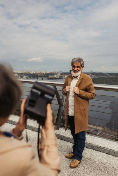 blurred woman holding vintage camera while taking photo of bearded senior husband on bridge near river - Fotografie, Obrázek