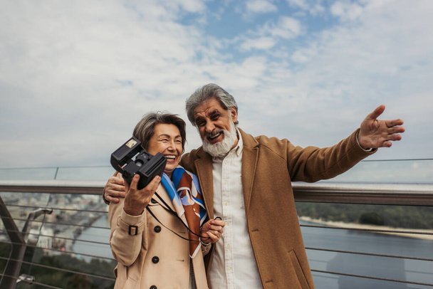 veselá starší žena drží vintage kameru, zatímco manžel usmívá a gestikuluje venku  - Fotografie, Obrázek
