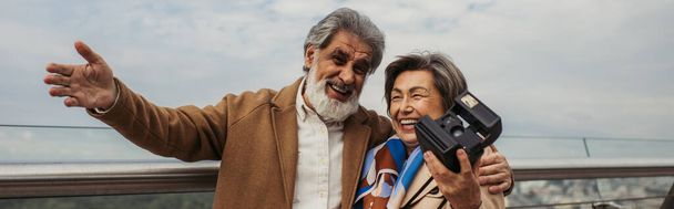 Χαρούμενη ηλικιωμένη γυναίκα κρατώντας vintage κάμερα, ενώ ο σύζυγος χαμογελά και gesturing έξω, πανό - Φωτογραφία, εικόνα