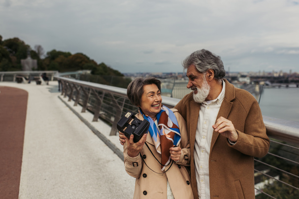 веселая пожилая женщина в плаще держит винтажную камеру и смотрит на счастливого мужа снаружи  - Фото, изображение